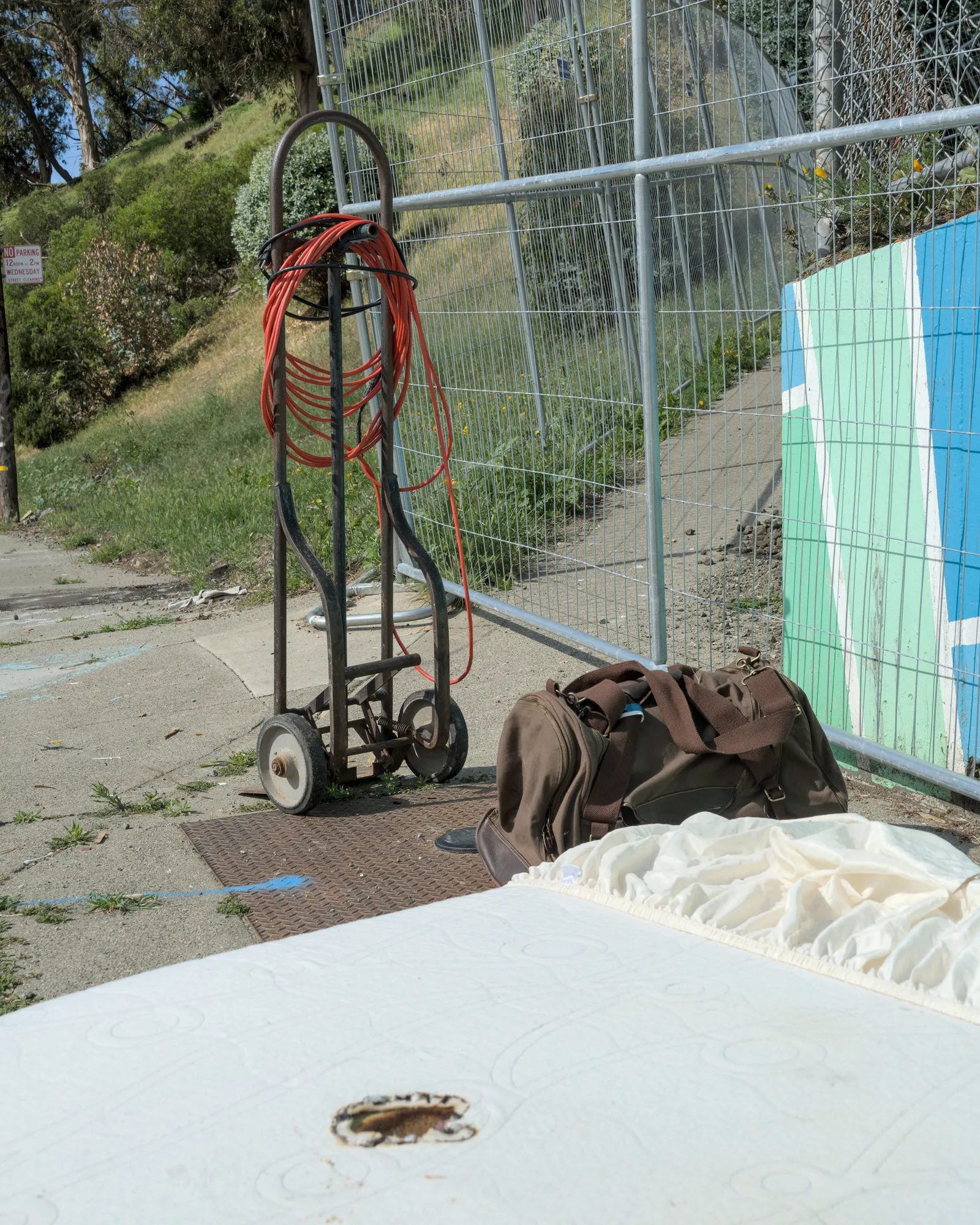 May 4th, 2026: A mattress with burn marks and a duffel bag lie on the sidewalk next to a fence.